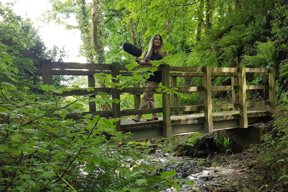 The artist Cheryl Beer stands on a wooden bridge over a river holding a sound recording microphone
