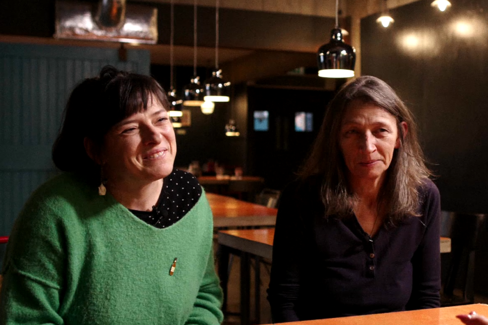 Two members of the band Kaolila are sat at a table in a dimly lit restaurant. The presenter Aleighcia Scott has their back to the camera and is interviewing Kaolila.