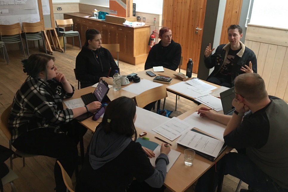 A group of people are sat around a square desk within a teaching room. One person is talking whilst the others listen, and hold pens to paper.
